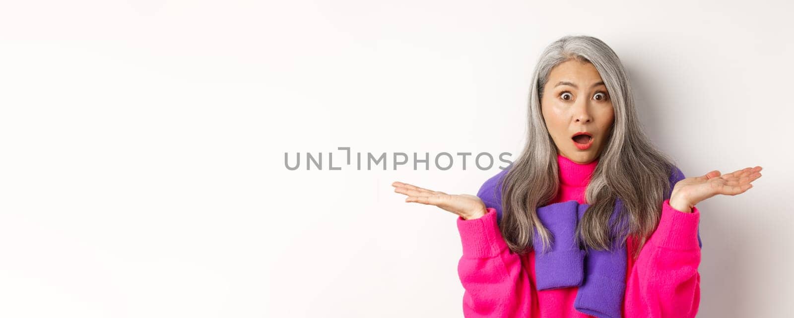 Close up of shocked asian mom staring at camera, spread hands sideways and gasping at camera confused, standing over white background.