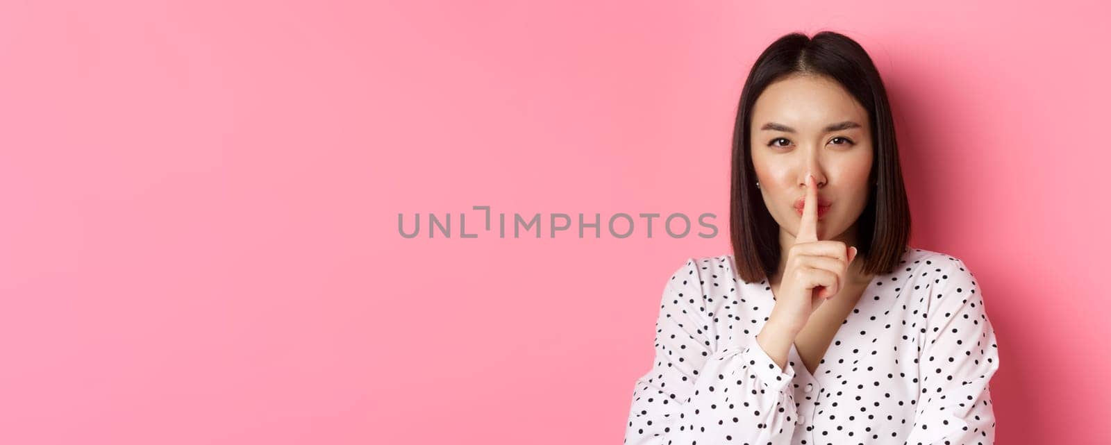 Close-up of mysterious asian woman hiding a secret, hushing and telling to keep quiet, standing over pink background by Benzoix