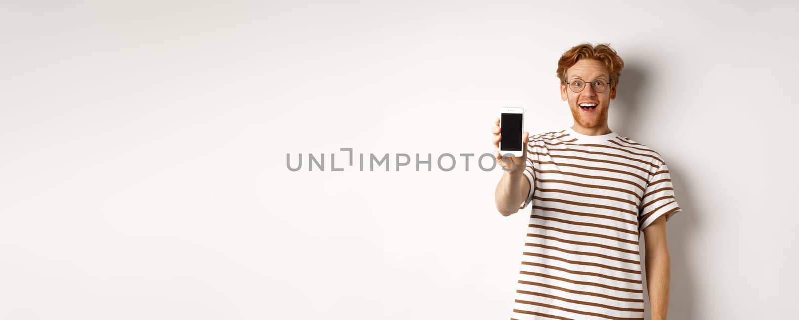 Technology and e-commerce concept. Happy young redhead man in glasses showing blank smartphone screen, looking at camera amazed, standing over white background.