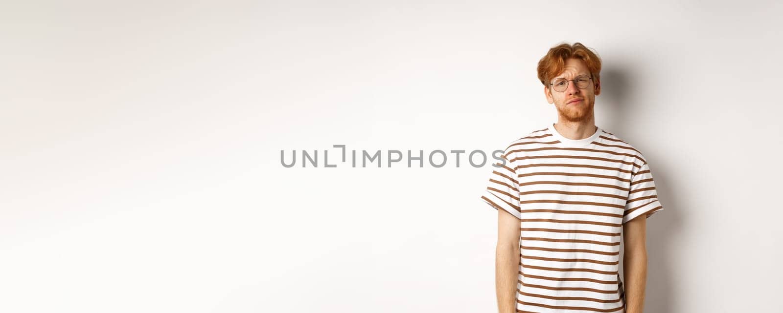 Reluctant and unamused redhead young man staring at camera, looking tired or displeased, standing over white background.