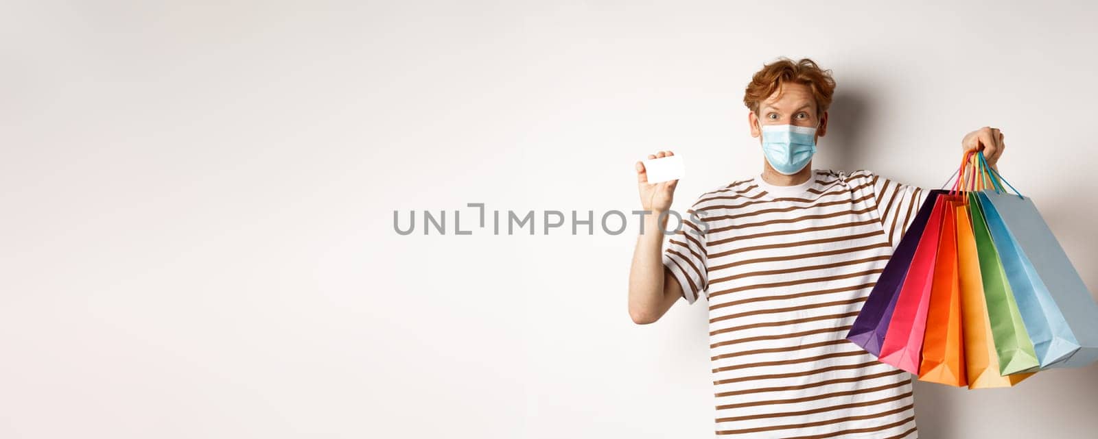 Social distancing, coronavirus concept. Young man with red hair, wearing face mask, showing shopping bags and plastic credit card, white background.