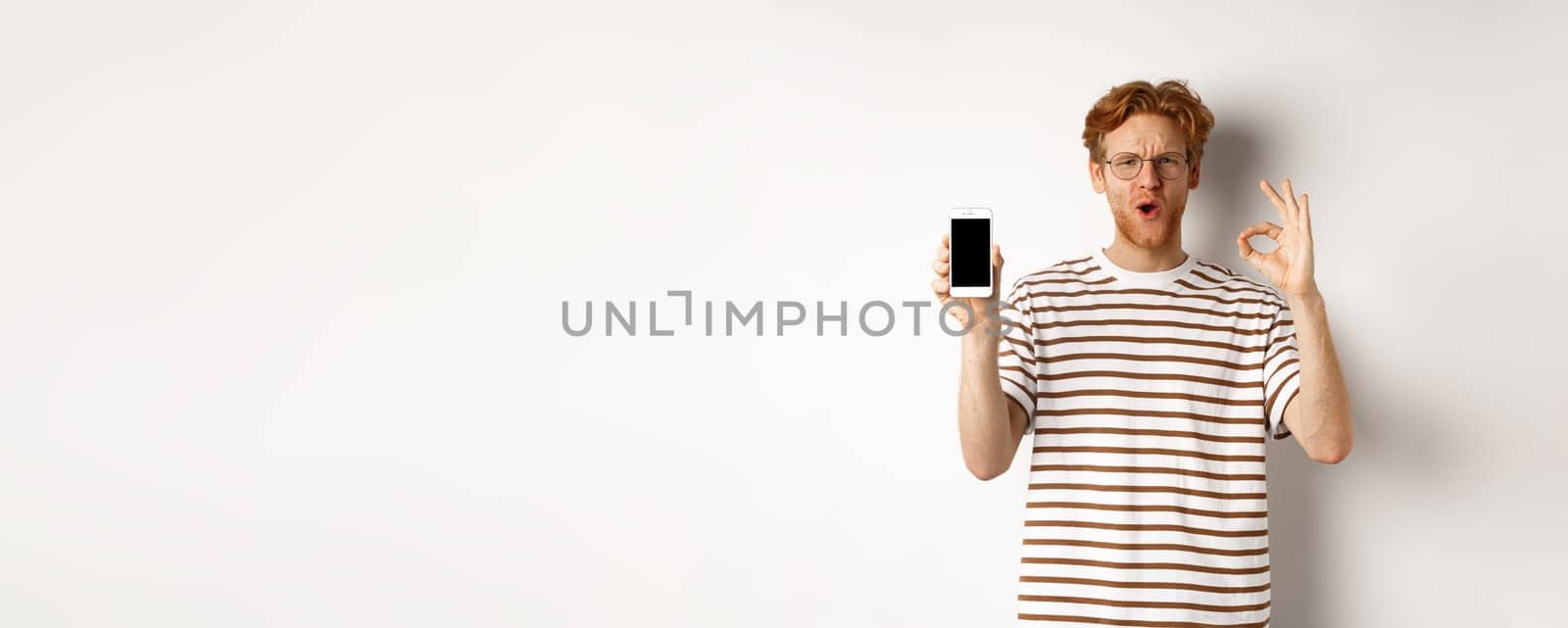 Technology and e-commerce concept. Young man with red hair showing okay sign and blank smartphone screen, praising awesome app or video game, white background.