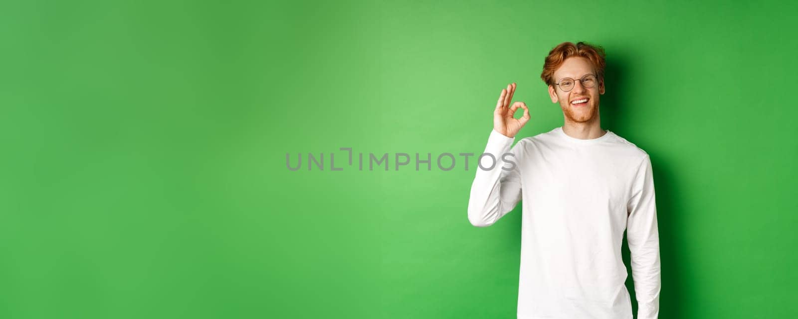 Happy male student with red hair, wearing glasses, showing okay sign in approval and smiling satisfied, standing over green background by Benzoix