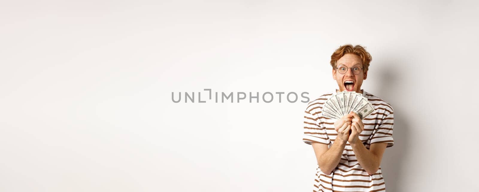 Lucky young man with red hair showing dollars, winning money and screaming of happiness, holding prize cash, standing over white background by Benzoix