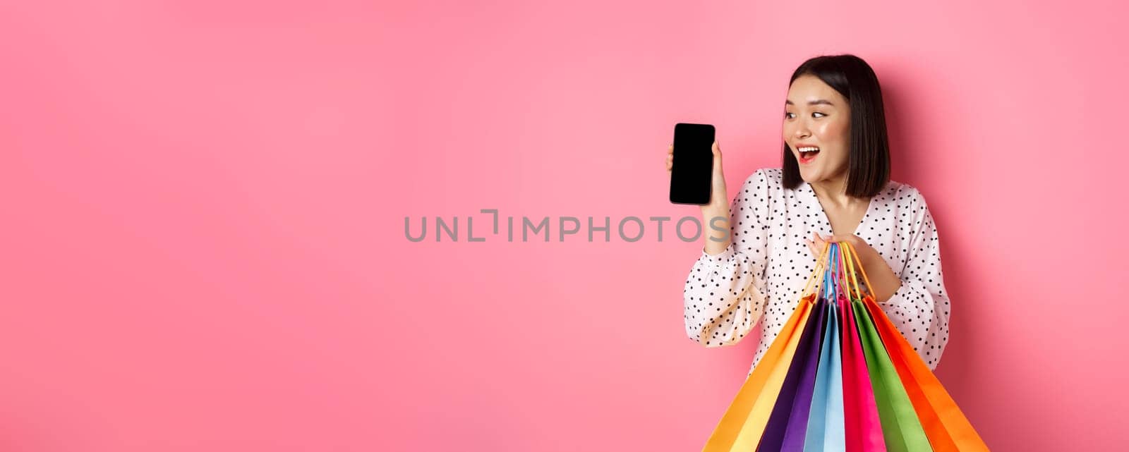 Attractive asian woman showing smartphone app and shopping bags, buying online via application, standing over pink background.