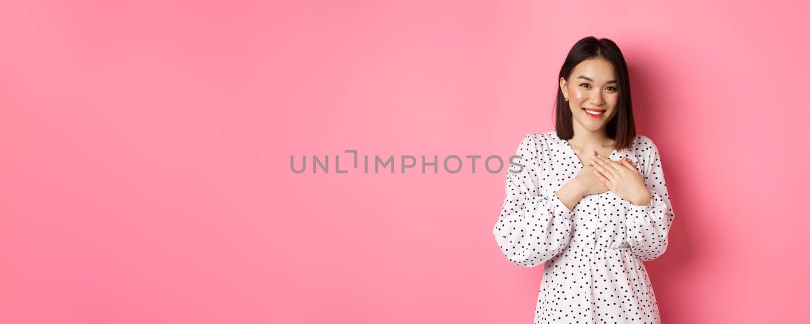 Thankful Korean girl in dress smiling, holding hands on heart and looking grateful at camera, touched with nice gesture, standing over pink background.