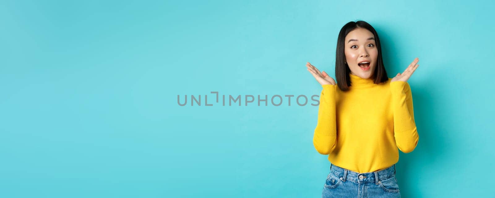 Beauty and fashion concept. Image of excited and surprised japanese girl saying wow with amazement, raising hands up near face, standing against blue background.