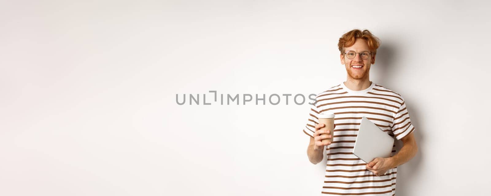 Handsome redhead male employee in glasses having break, drinking coffee and holding laptop, standing over white background.