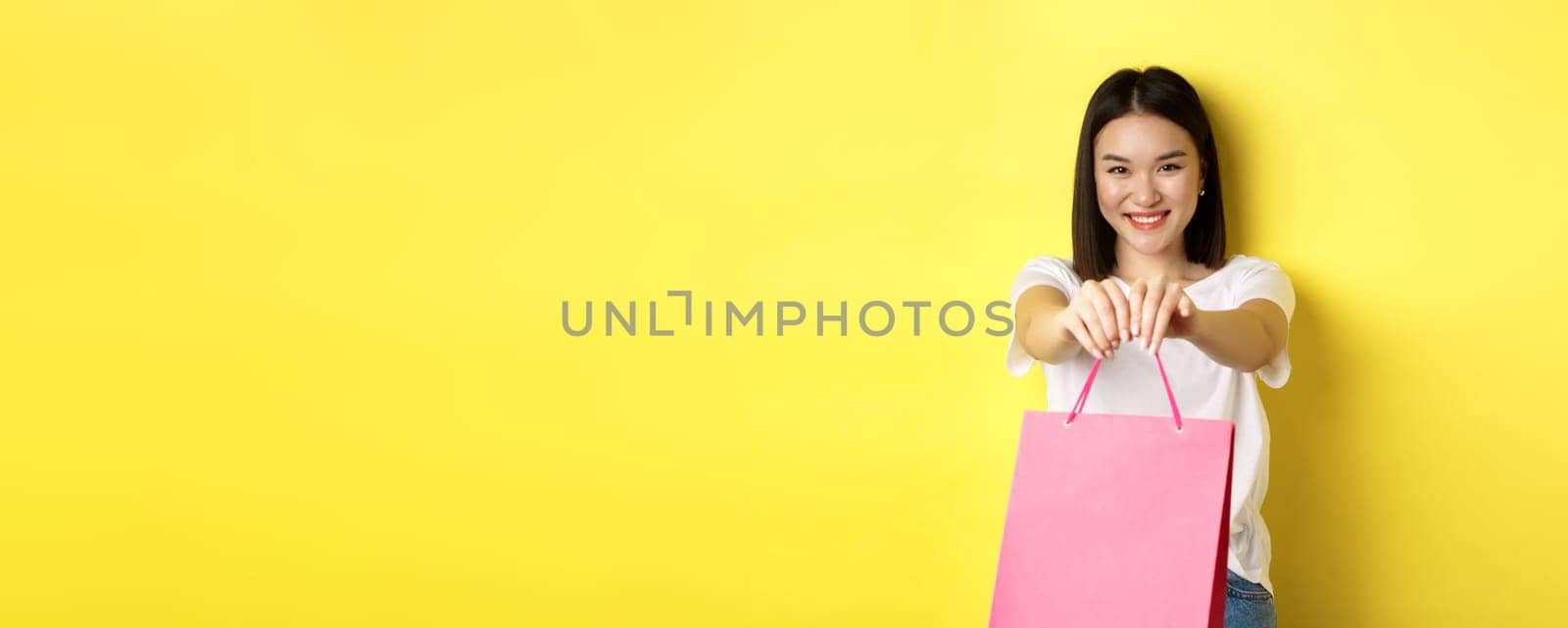 Cute asian girl giving you gift, stretch out hand with pink shopping bag and smiling, congratulating with holiday, standing over yellow background by Benzoix