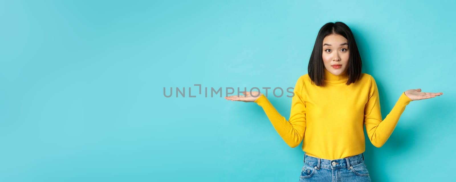 Image of indecisive asian woman shrugging shoulders, spread hands sideways and looking clueless at camera, standing indecisive against blue background.