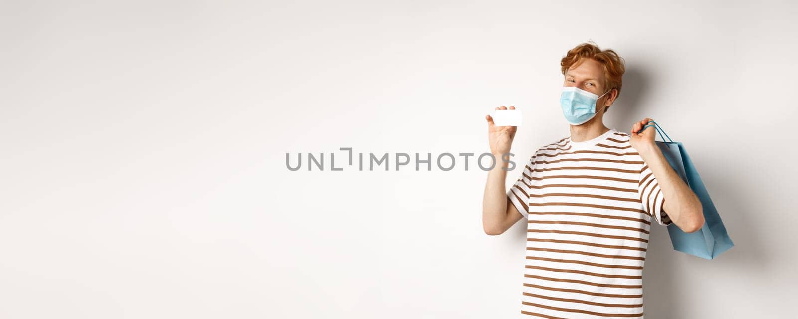 Concept of covid-19 and lifestyle. Happy young shopper in face mask holding shopping bag and showing plastic credit card, buying with discounts, white background.