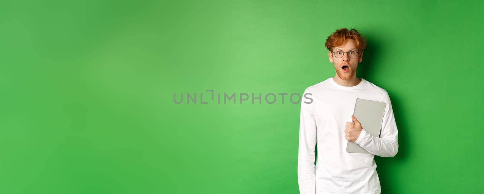 Surprised young man holding laptop and staring at camera, wearing glasses and white t-shirt, standing over green background.