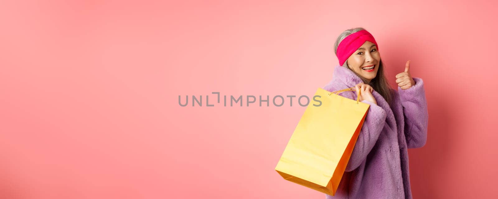Stylish asian senior woman going shopping, carry paper bag on shoulder and showing thumb-up, recommending store discounts, pink background by Benzoix