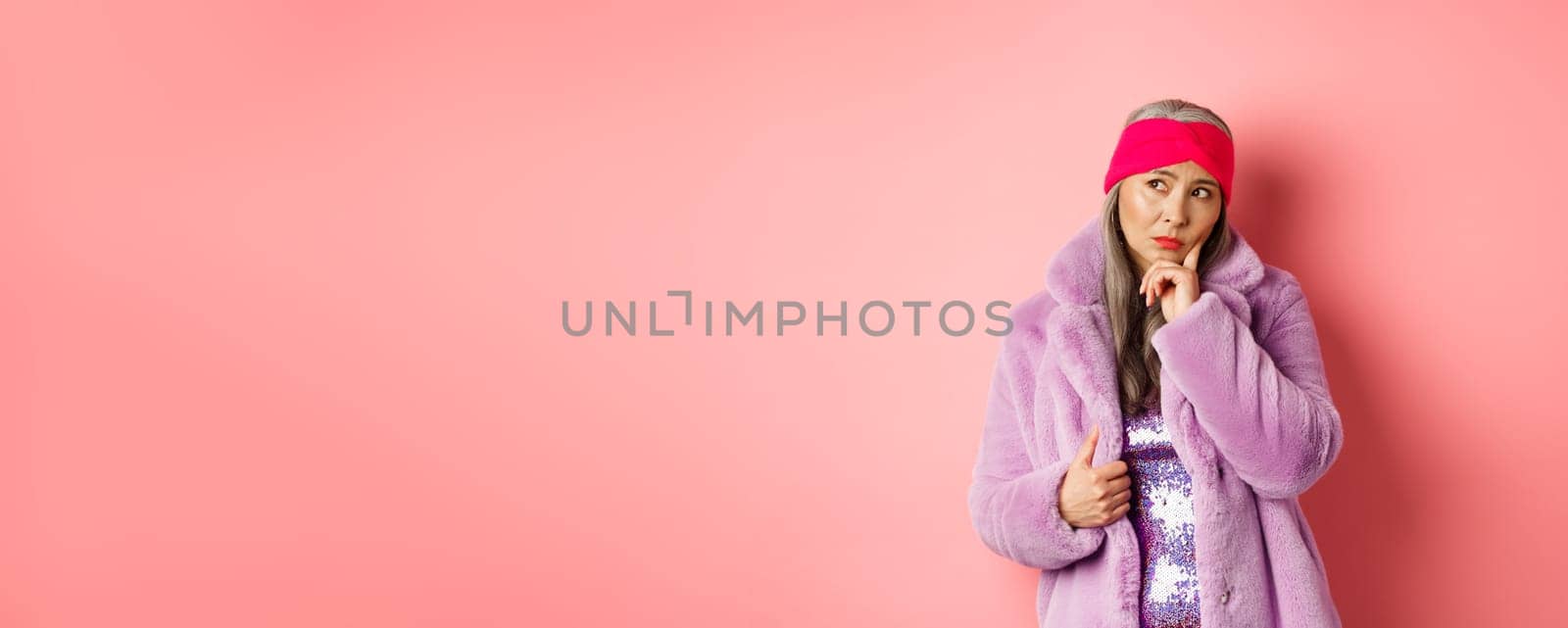 Fashionable asian senior woman looking doubtful, thinking and frowning indecisive, standing in purple faux fur coat and looking left, standing over pink background.