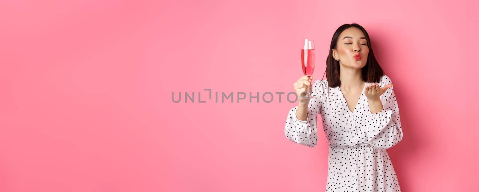 Romantic asian woman raising glass of champagne and sending air kiss at camera, celebrating and having fun, standing over pink background.