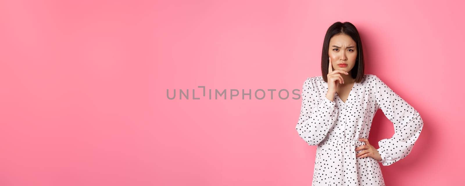 Concerned asian woman staring at camera suspicious, having doubts, thinking about something with serious face, standing against pink background by Benzoix