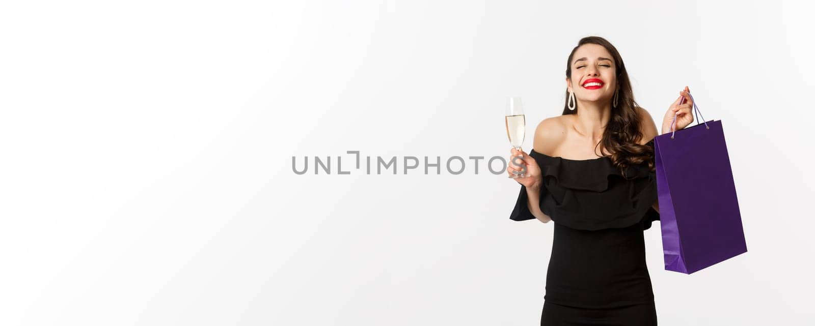 Happy smiling woman celebrating, holding present in shopping bag and glass of champagne, standing in black dress over white background by Benzoix