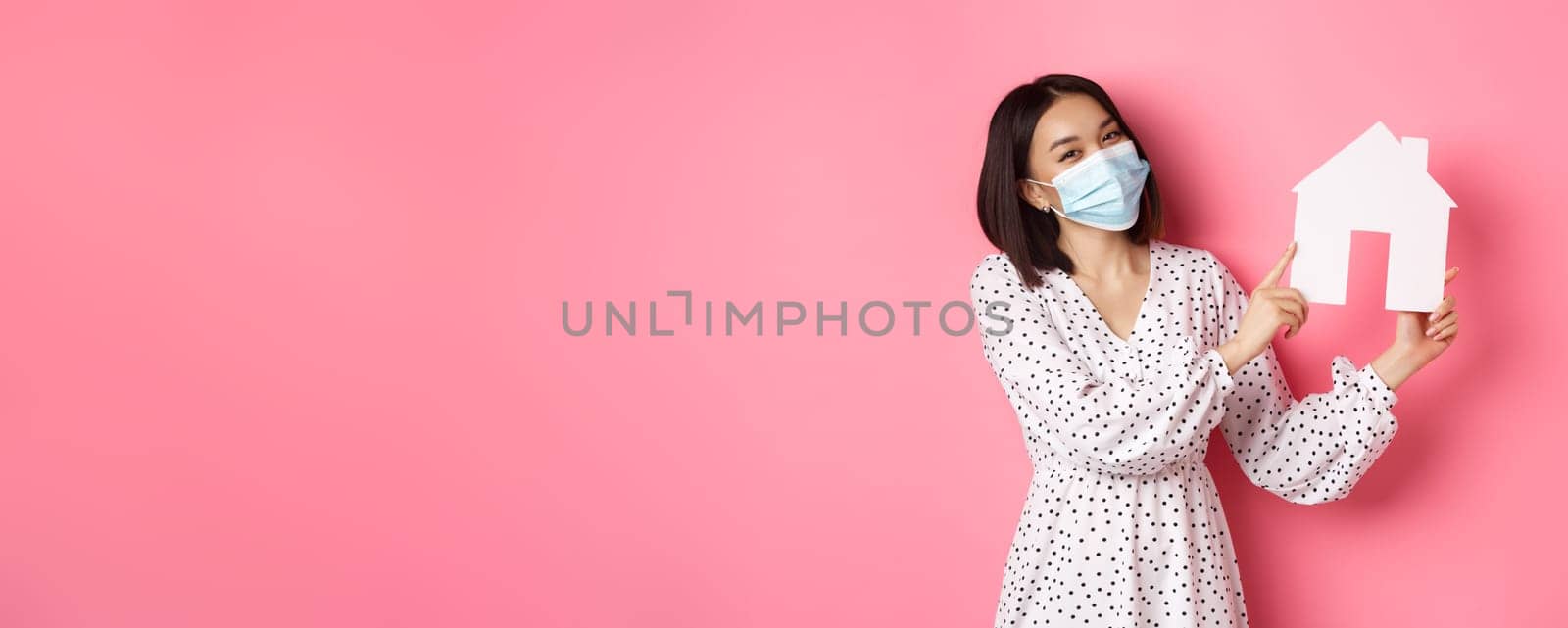 Covid-19, real estate and lifestyle concept. Cute asian woman in face mask selling houses, showing model of home and looking at camera, standing over pink background.