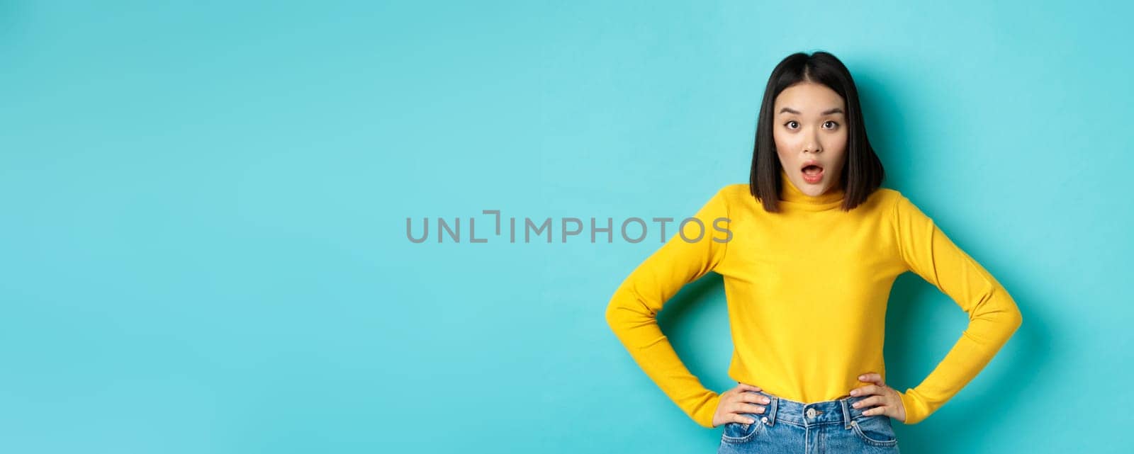 Portrait of surprised asian woman drop jaw, listening to big news, looking amazed at camera, standing in yellow sweater against blue background by Benzoix