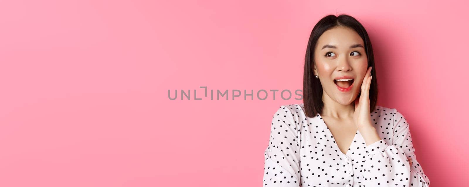 Coquettish asian woman in blushing, touching cheek and looking left amused, standing over pink background.