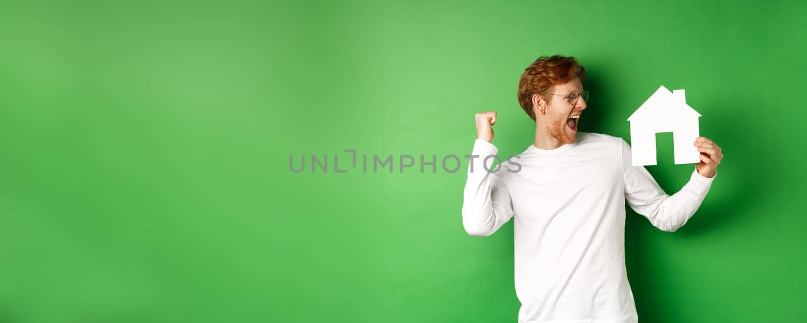Real estate. Cheerful redhead man in glasses celebrating, buying a house, showing paper home cutout and scream with joy, standing over green background.