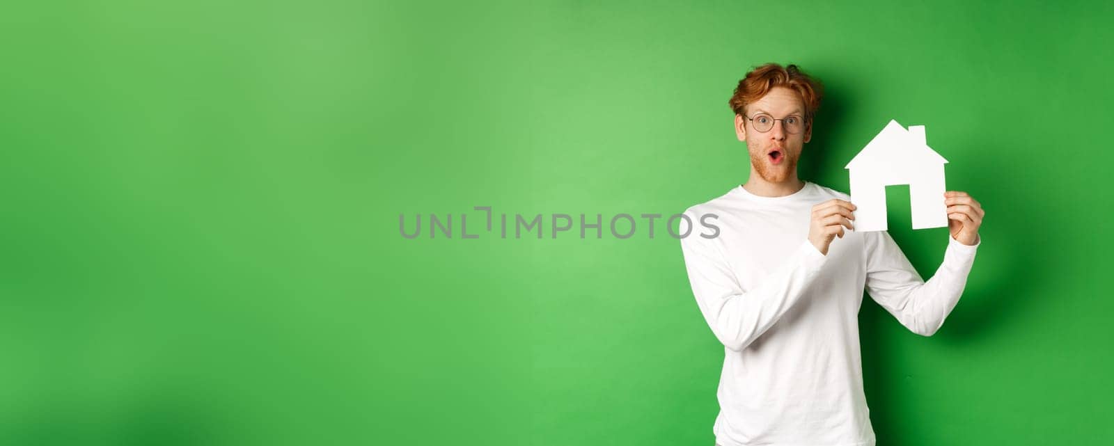 Real estate and buying property concept. Surprised young redhead man showing paper house model and looking amazed, standing over green background by Benzoix