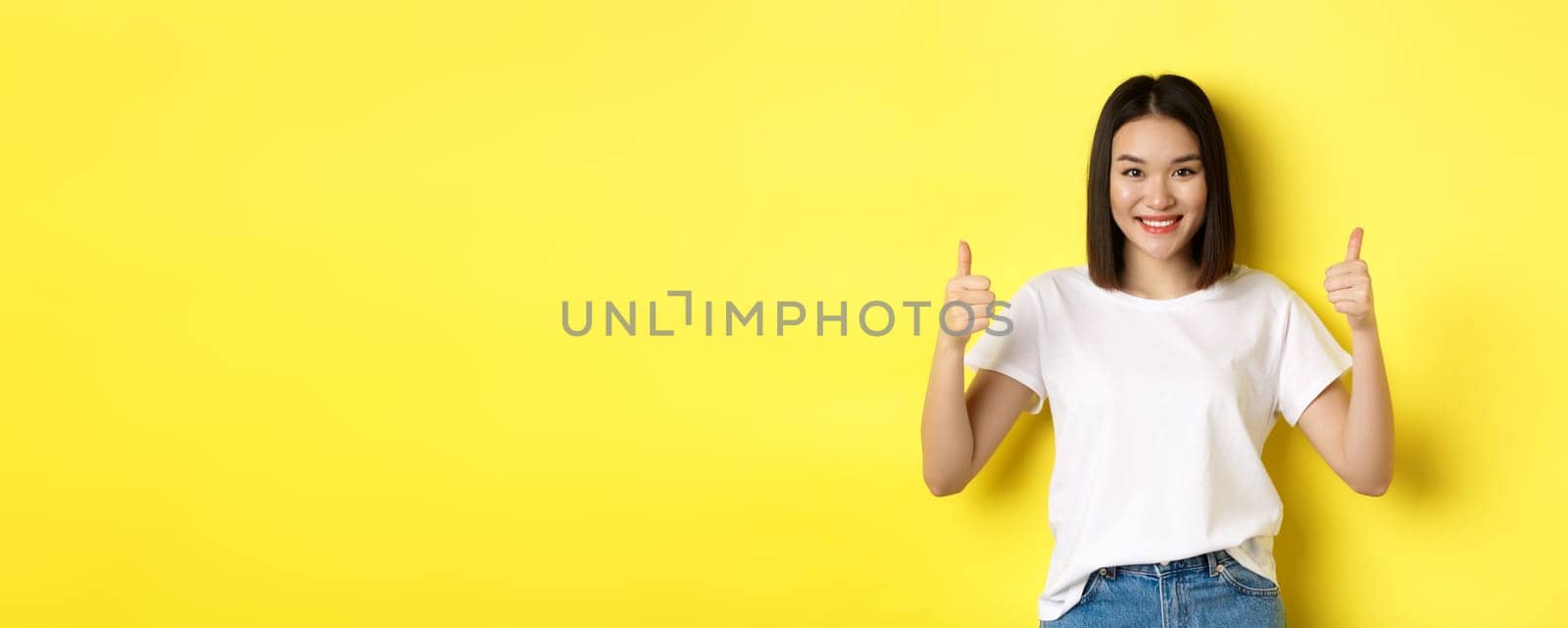 Pretty young asian woman in white t-shirt, showing thumbs up and smiling, praise good offer, recommend product, standing satisfied over yellow background.