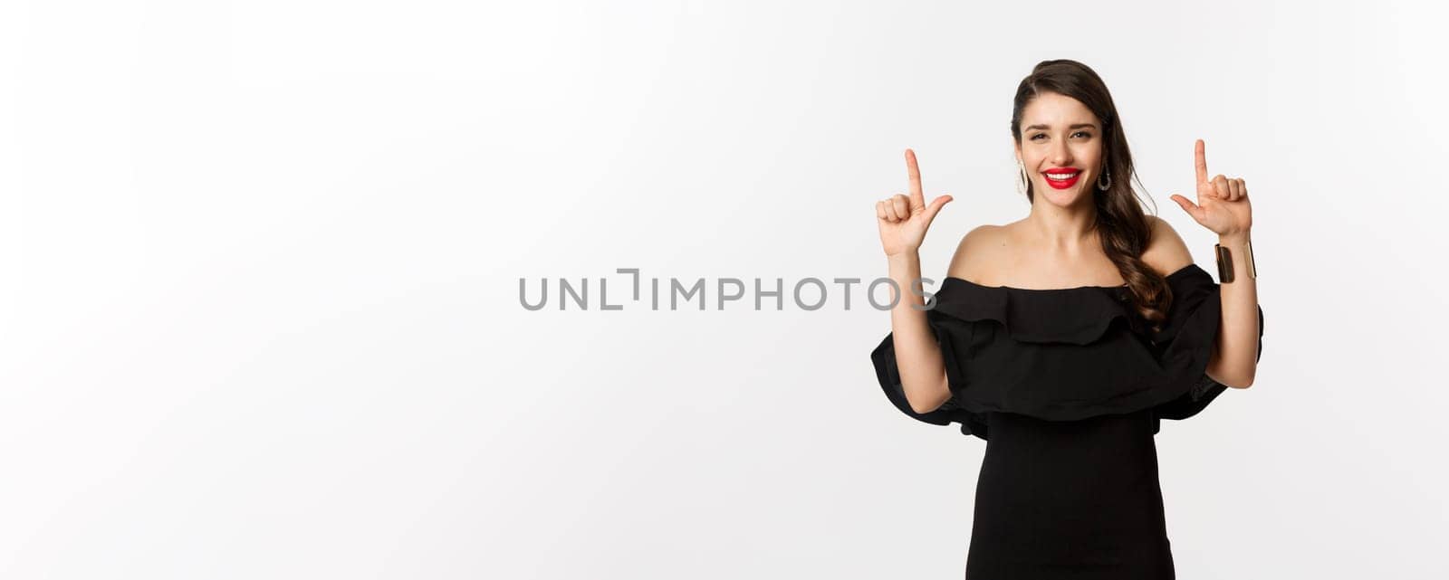 Fashion and beauty. Charming woman with red lips, black dress, smiling happy and pointing fingers up, showing logo, white background.