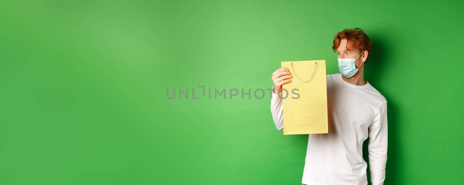 Surprised redhead guy receiving a present, looking at shopping bag with curious face, standing over green background in face mask from covid-19.