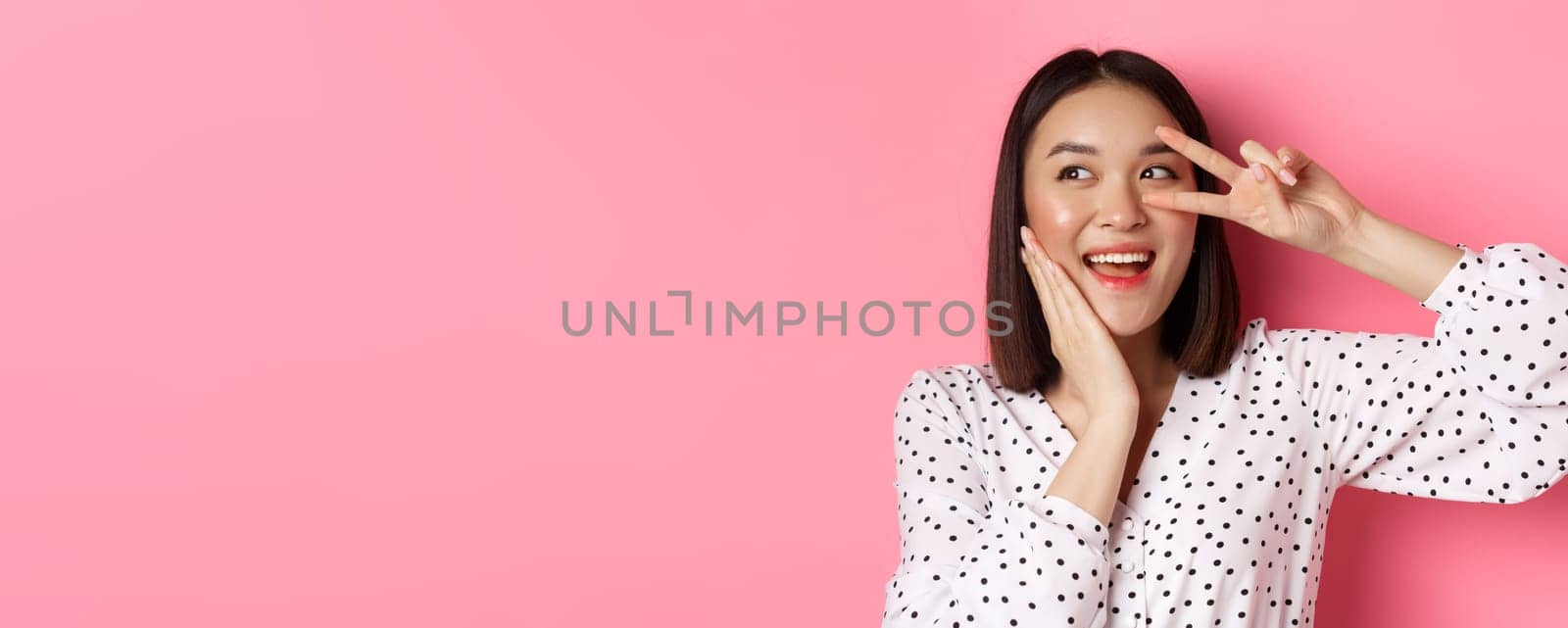 Beauty and lifestyle concept. Close-up of pretty asian woman showing peace sign and touching cheek, smiling happy at camera, standing over pink background.