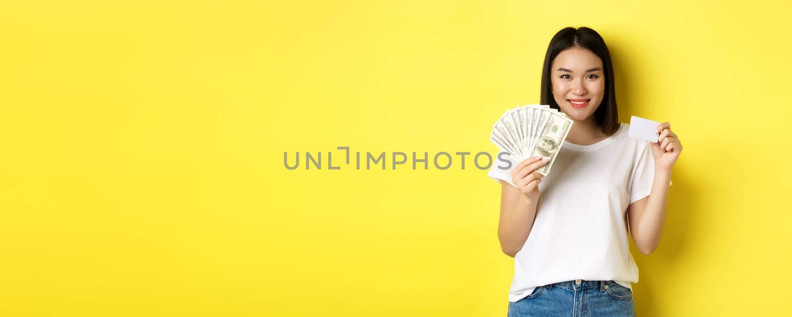 Beautiful asian woman with short dark hair, wearing white t-shirt, showing money in dollars and plastic credit card, standing over yellow background by Benzoix