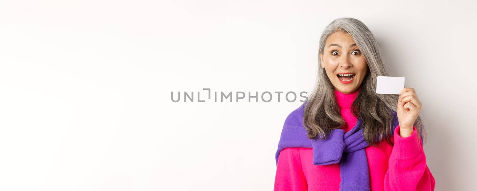 Shopping concept. Happy asian old aldy looking impressed and showing plastic credit card of her bank, standing over white background by Benzoix
