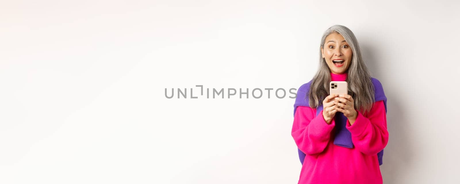 Surprised asian woman smiling at camera after reading promotion on smartphone, standing with mobile phone over white background.