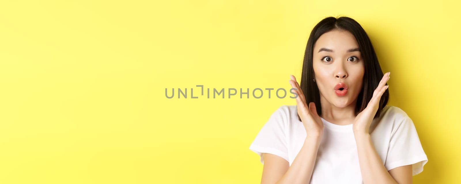 Close up of surprised asian girl say wow, stare at camera amzed with hands near face, standing over yellow background.