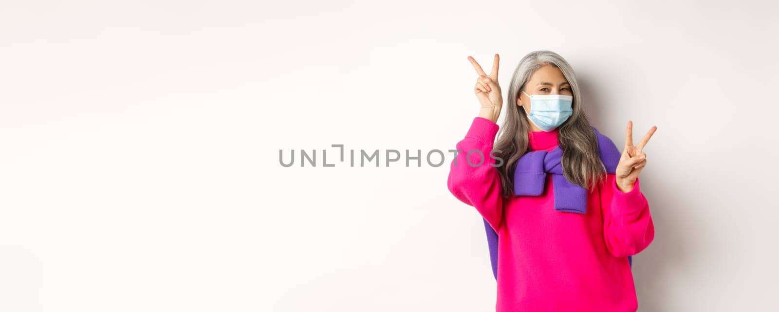 Covid, pandemic and social distancing concept. Cheerful and stylish asian senior woman wearing medical mask and showing peace signs, standing over white background by Benzoix