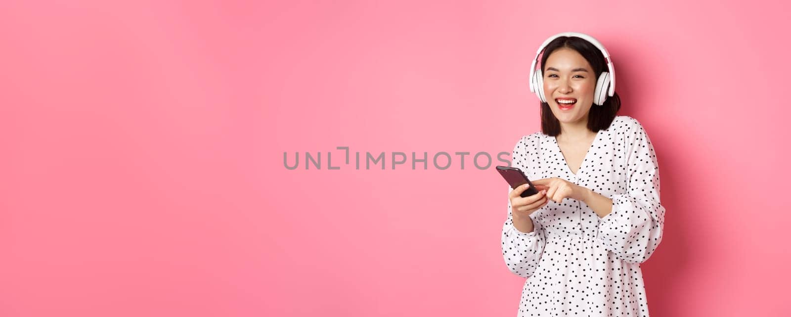 Beautiful asian woman listening music in headphones, using mobile phone, smiling happy at camera, standing over pink background.