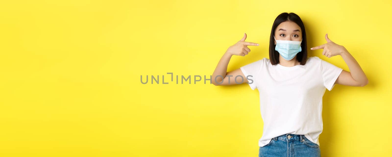 Covid-19, quarantine and social distancing concept. Young asian woman in white t-shirt, pointing at her medical mask from coronavirus, yellow background.