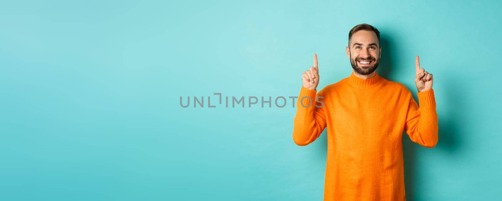 Satisfied and happy buyer pointing fingers up, smiling pleased, standing in orange sweater against turquoise background.