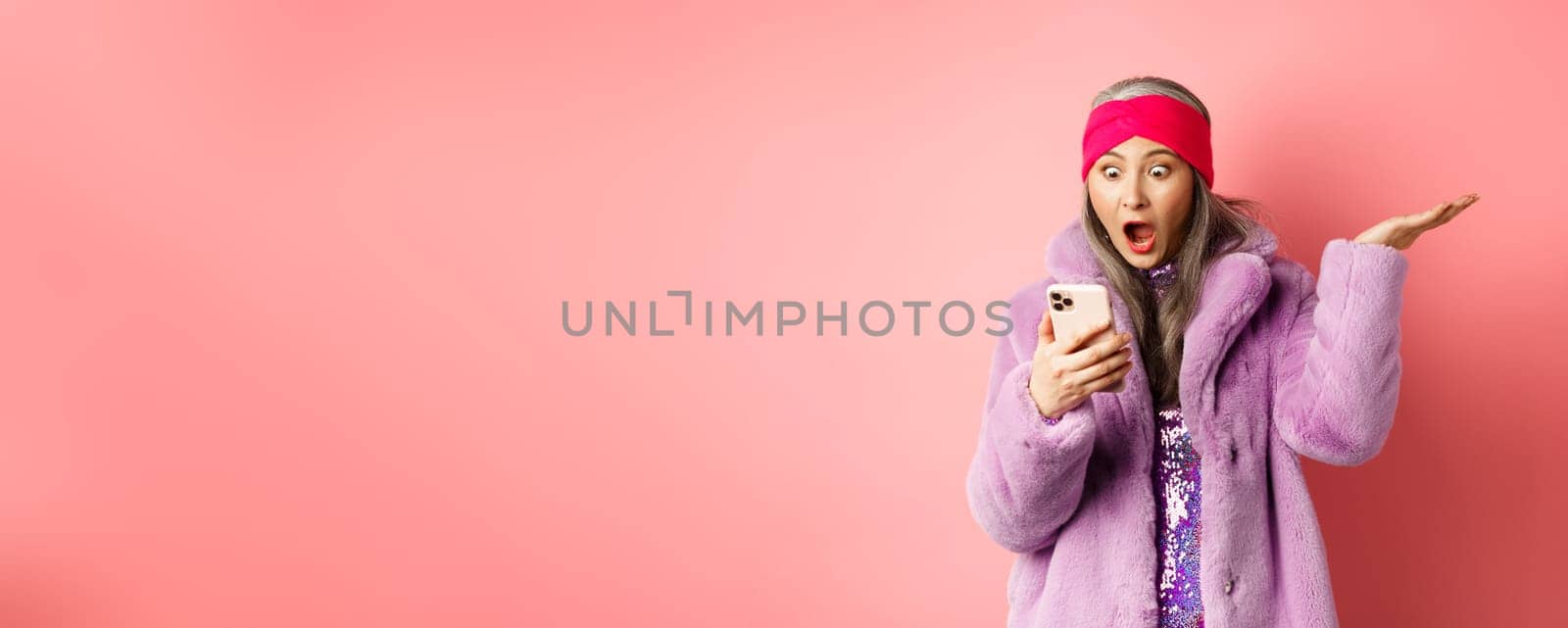 Online shopping and fashion concept. Amazed and surprised asian senior woman looking at smartphone screen, reading on mobile phone and gasping, pink background by Benzoix