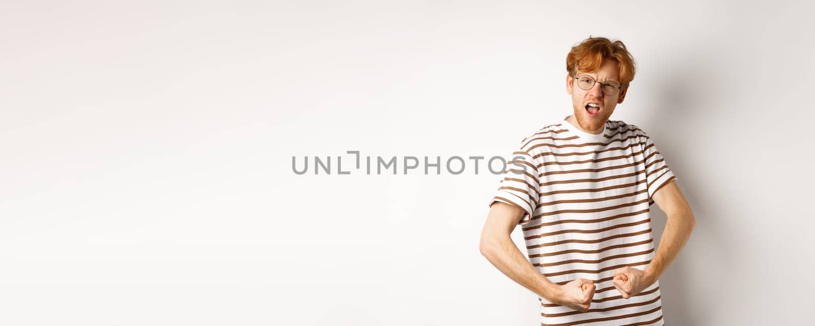 Image of confident and strong redhead man flexing biceps, showing muscles after gym, standing over white background by Benzoix