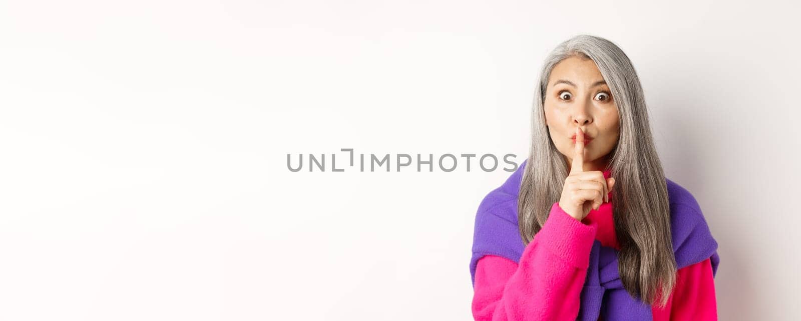 Close-up of stylish senior asian woman in hipster outfit telling hush, shushing at looking at camera, show taboo gesture, standing over white background.