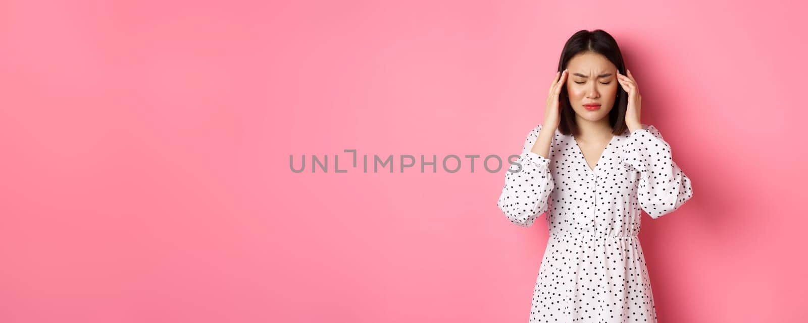 Image of upset asian woman having headache, feeling unwell or dizzy, close eyes and massaging head, suffering migraine, standing over pink background by Benzoix