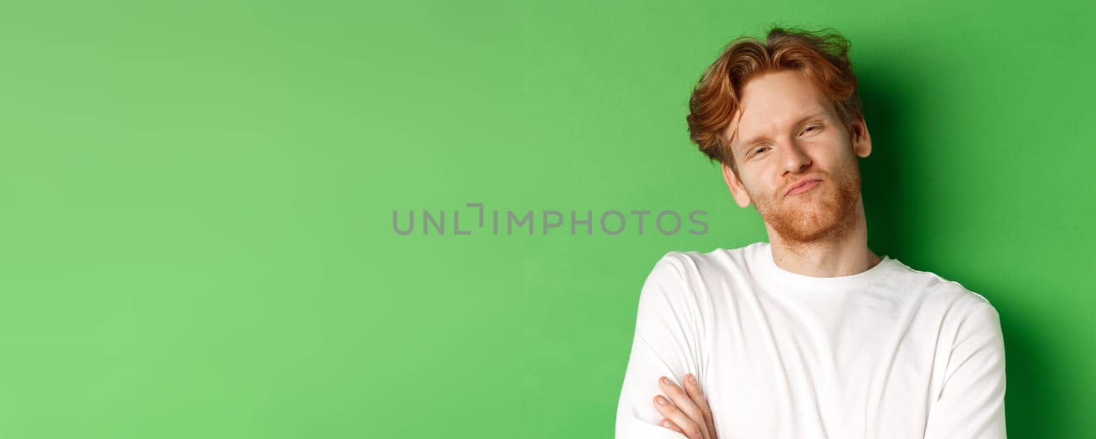 Emotions and fashion concept. Close-up of skeptical young man with red hair and beard pucker lips, looking at camera as waiting for something, standing over green background.