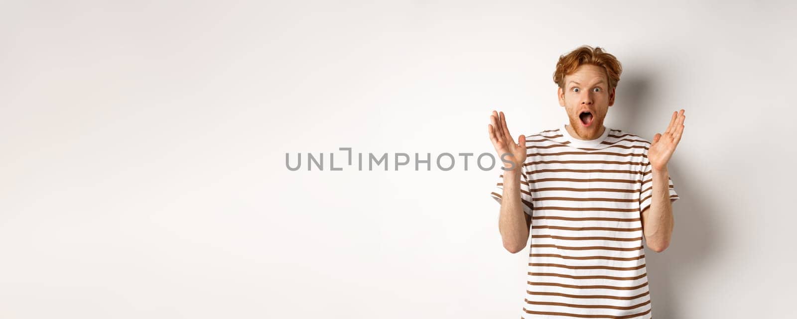 Surprised redhead man scream of amazement, staring with disbelief and excitement, hear awesome news, standing over white background.