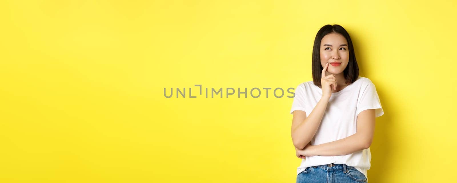 Beauty and fashion concept. Pensive asian woman thinking, looking thoughtful while pondering something, standing over yellow background by Benzoix