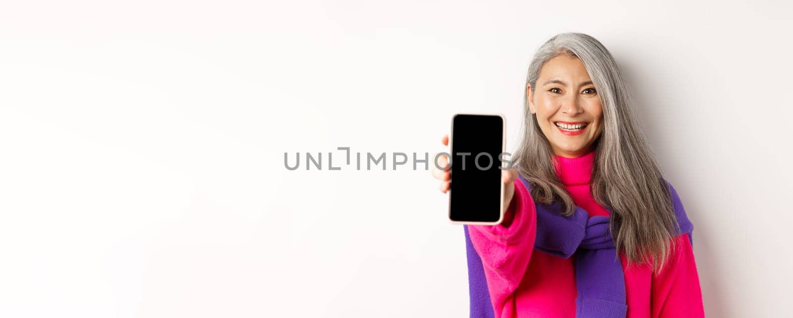 Online shopping. Close up of modern asian senior woman extending hand with mobile phone, showing blank smartphone screen and smiling, standing over white background by Benzoix