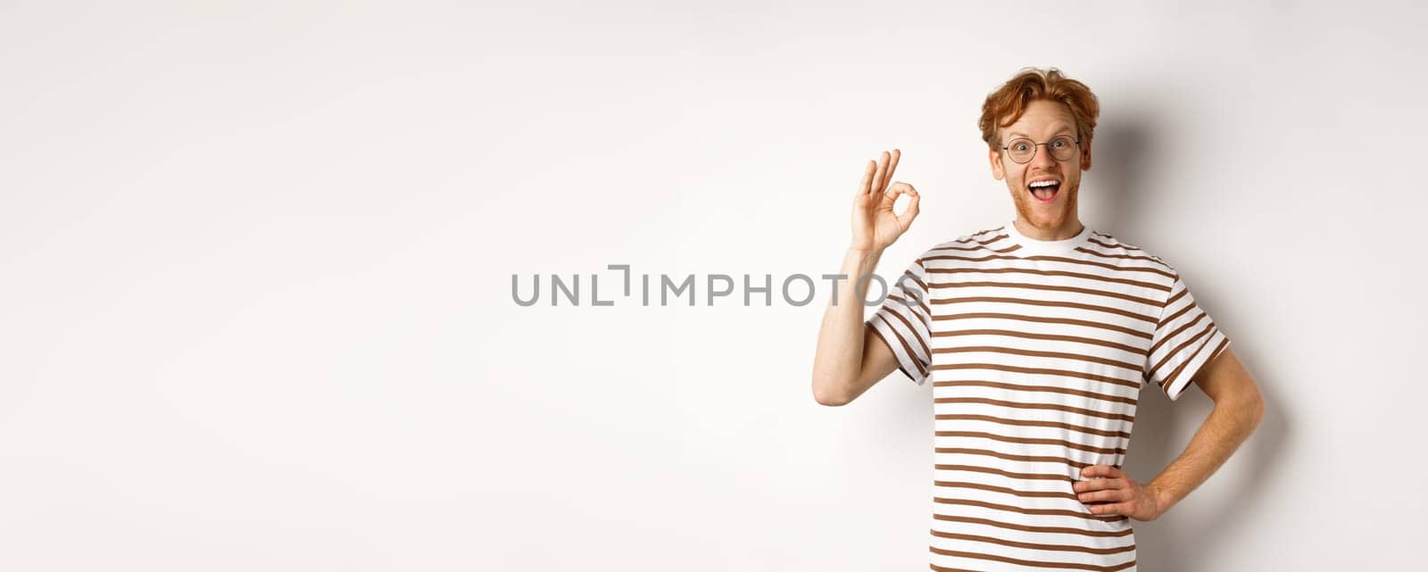 Excellent job. Happy redhead man praising you, showing OK sign and smiling amazed, approve and like something good, standing over white background.
