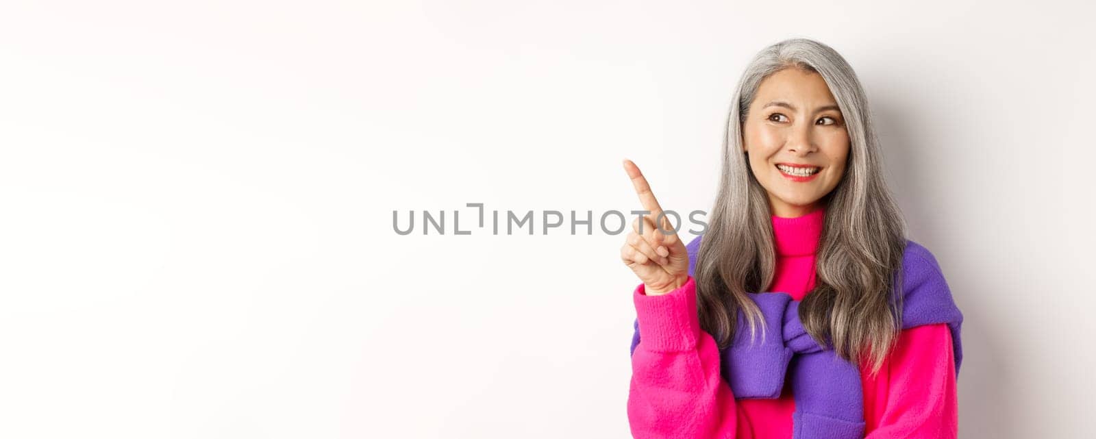 Close-up of beautiful asian senior woman smiling, looking and pointing upper left corner advertisement, checking out promo offer, standing over white background.