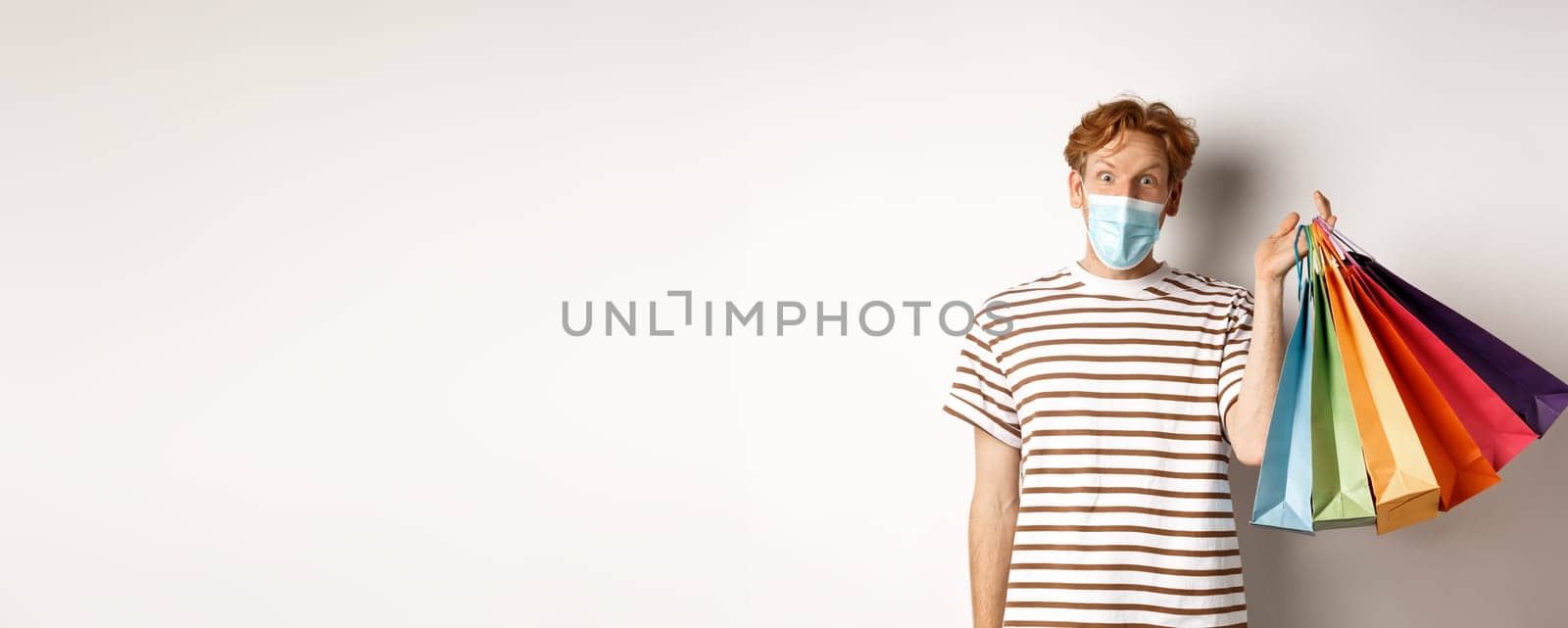 Pandemic and lifestyle concept. Handsome young man in face mask amazed with special discounts, holding shopping bags and staring excited, white background.