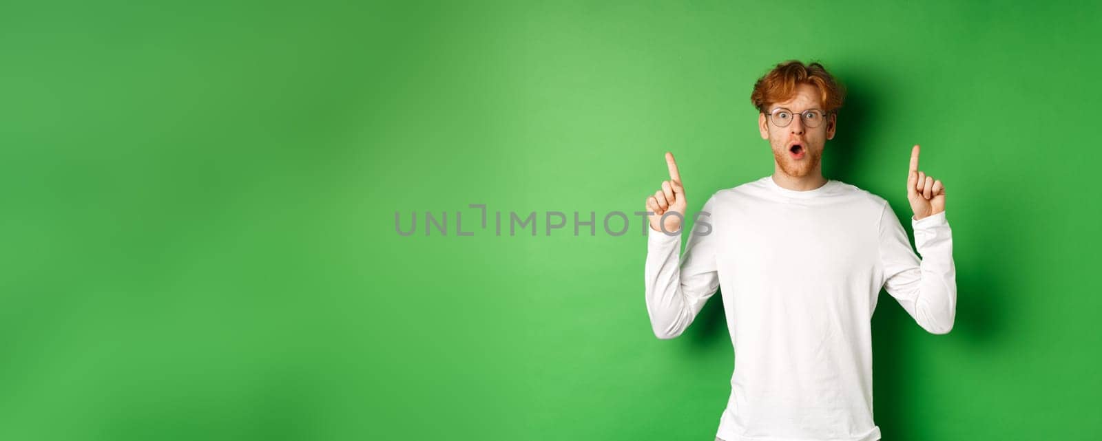 Shocked and worried redhead man in glasses gasping and staring at camera, pointing fingers up at advertisement, standing over green background.